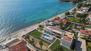 An aerial view of a coastal area with a clear blue sea, a beach, and a residential area with modern buildings and a swimming pool. - Amyntas Seafront Hotel - Adults Only (Sithonia)