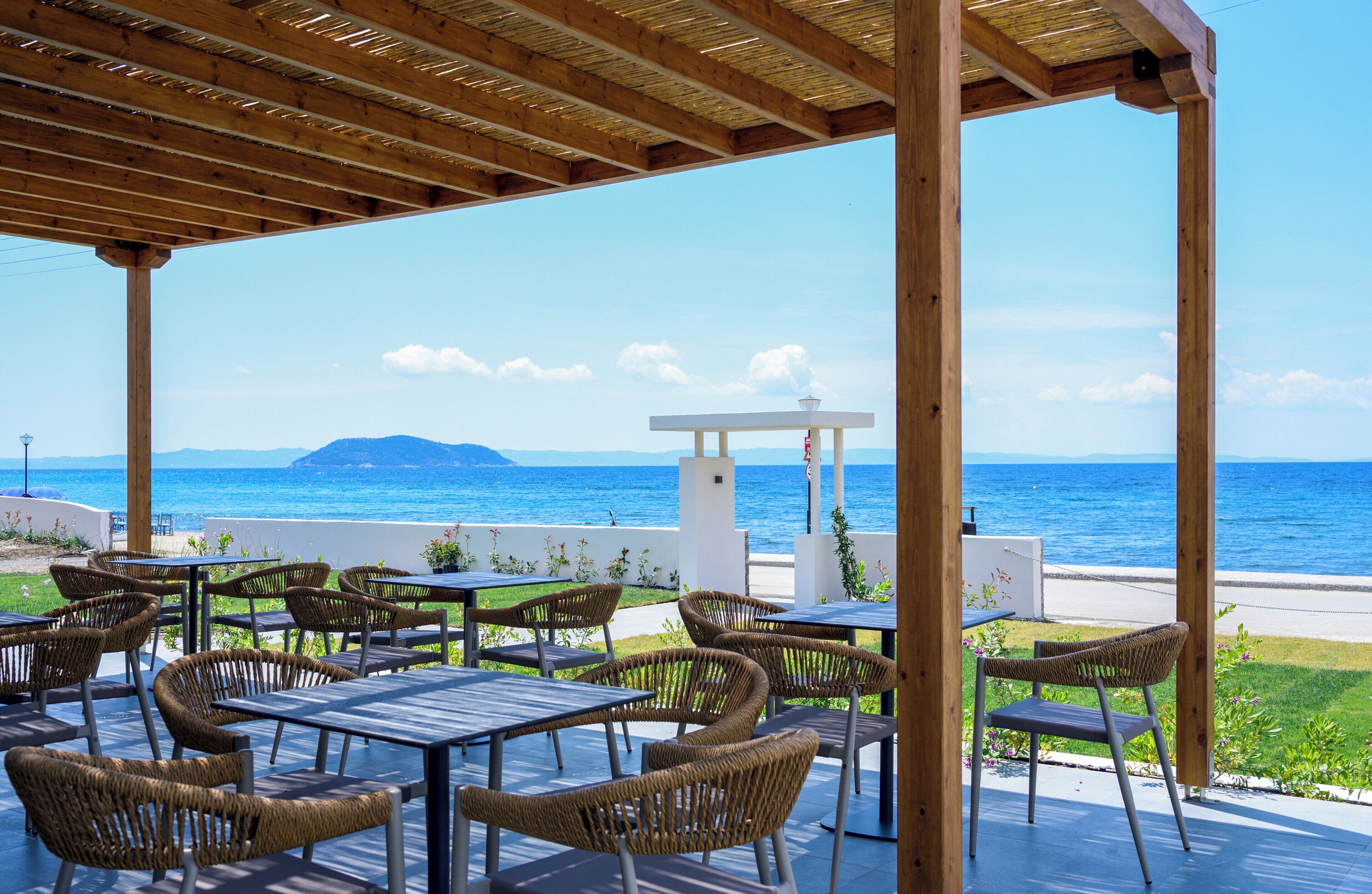 A covered outdoor seating area with tables and wicker chairs, overlooking a beach and the sea.