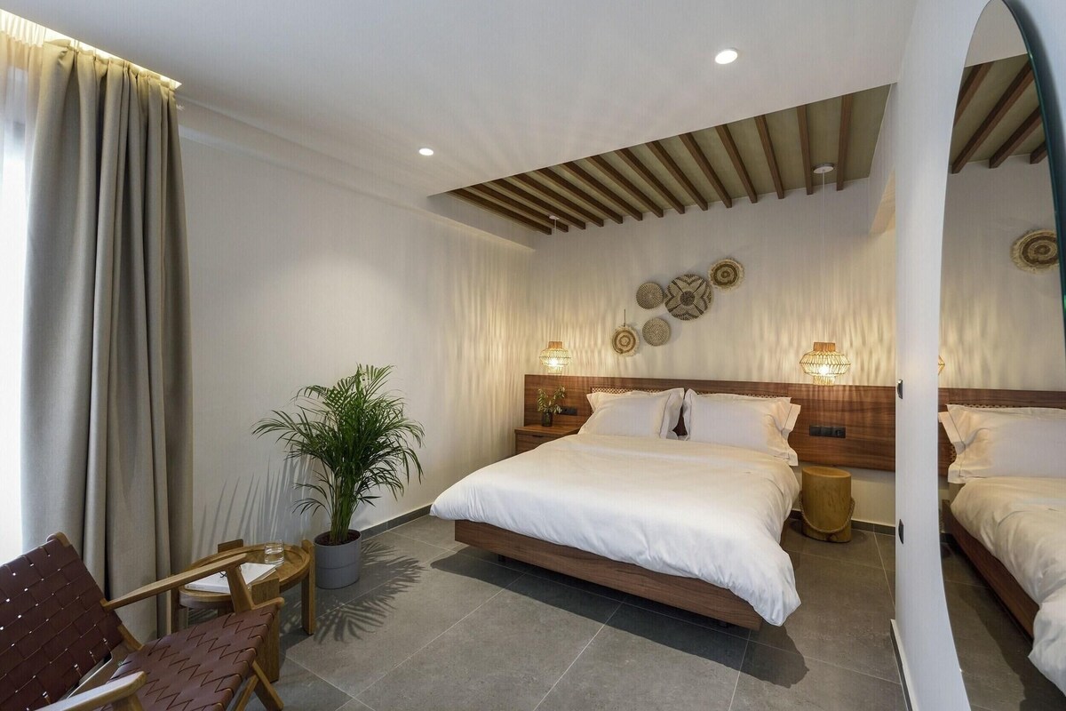 Una habitación de hotel moderna con una cama grande, dos mesitas de noche, una planta en maceta y una zona de estar con sillas.