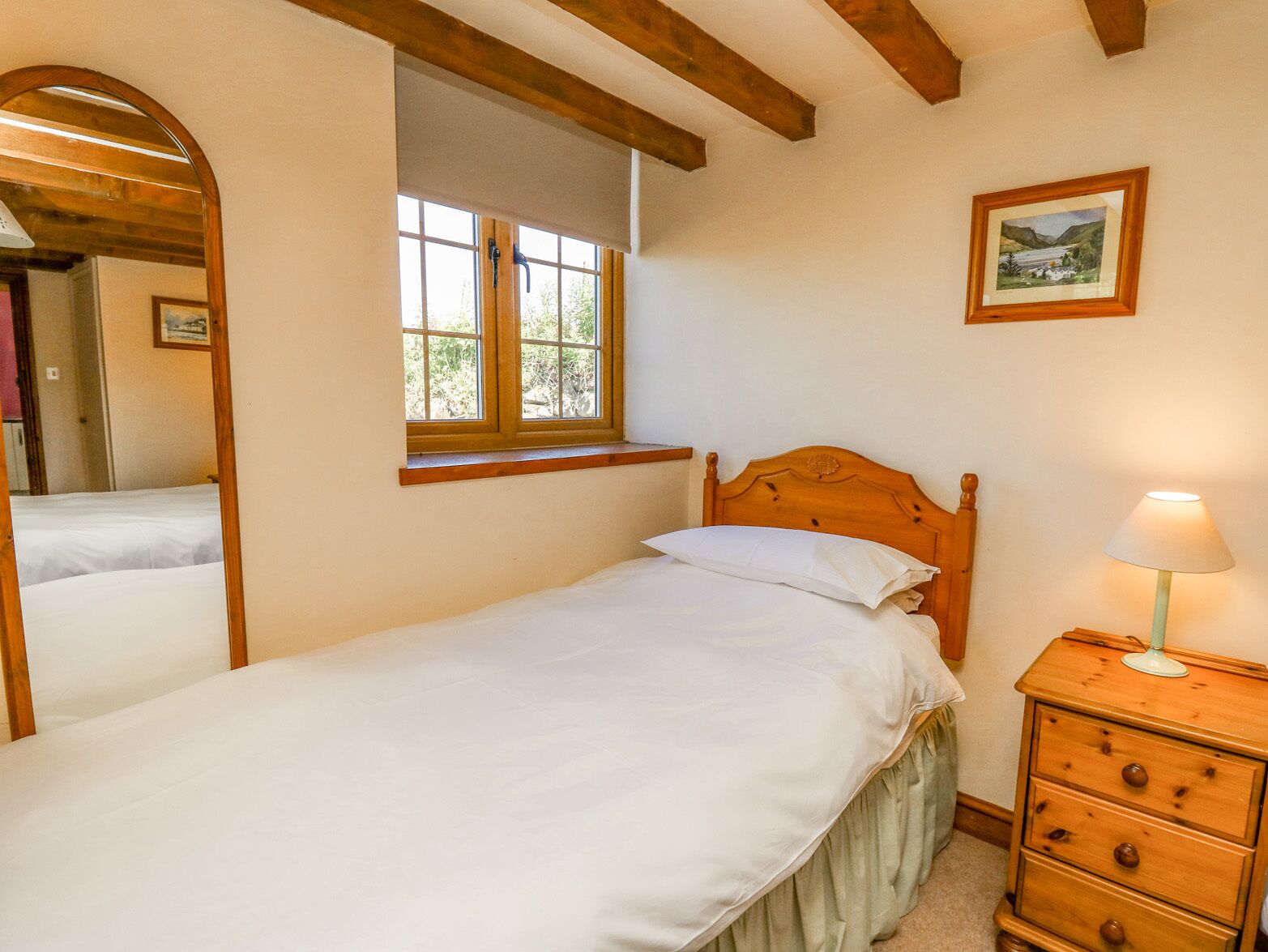 A bedroom with a single bed, a wooden headboard, a window with a view, a mirror, a dresser, and a lamp.