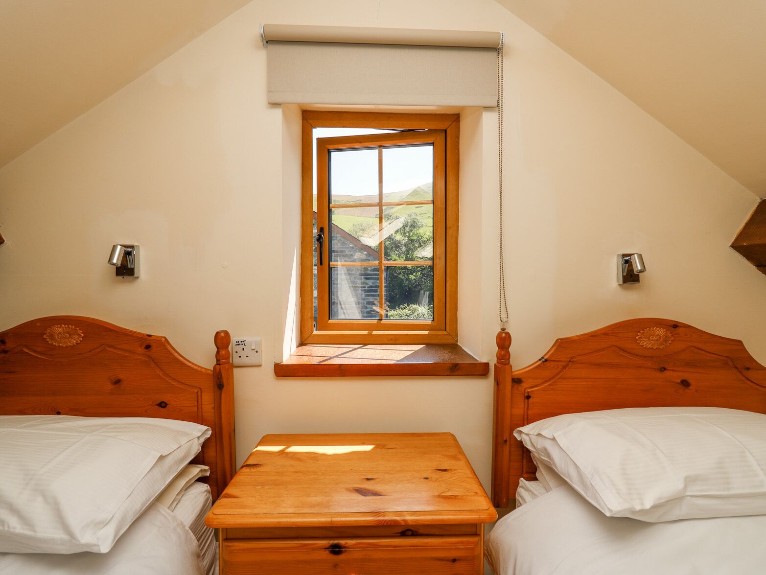 A small, cozy room with two wooden beds, a wooden table, and a window with a view of a green landscape.