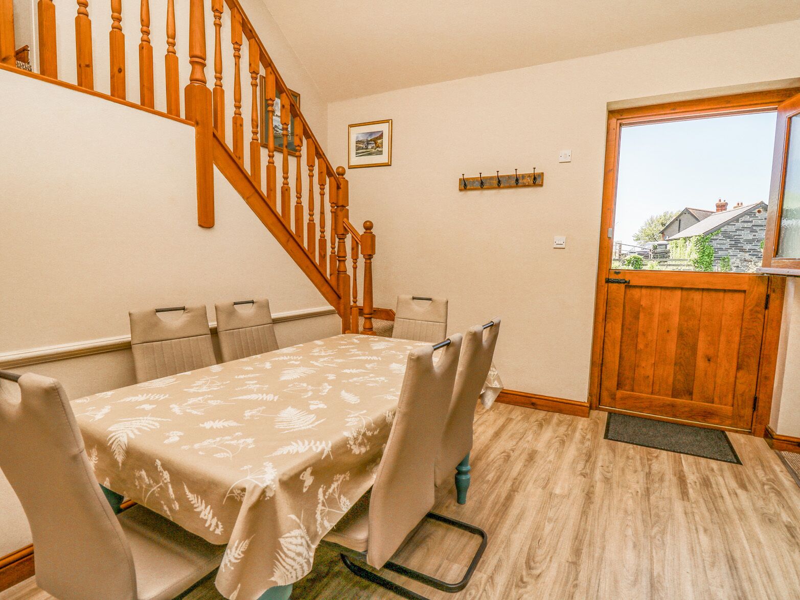 A dining area with a table set for four, wooden stairs, a coat rack, and a window with a view of houses.