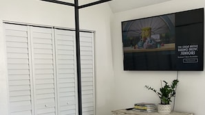 Un dormitorio con cama con dosel, un armario blanco, un aparador con adornos y un televisor en la pared que muestra un programa.