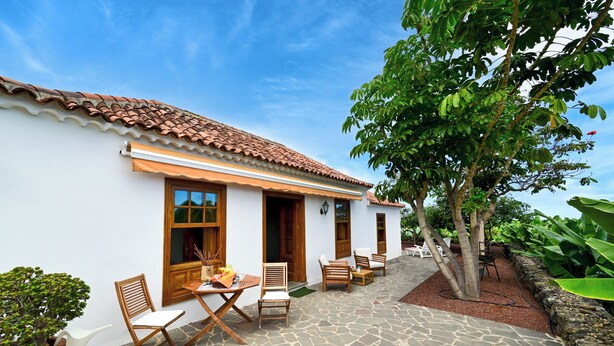 A courtyard with stone flooring, wooden furniture, and a white building with a tiled roof.