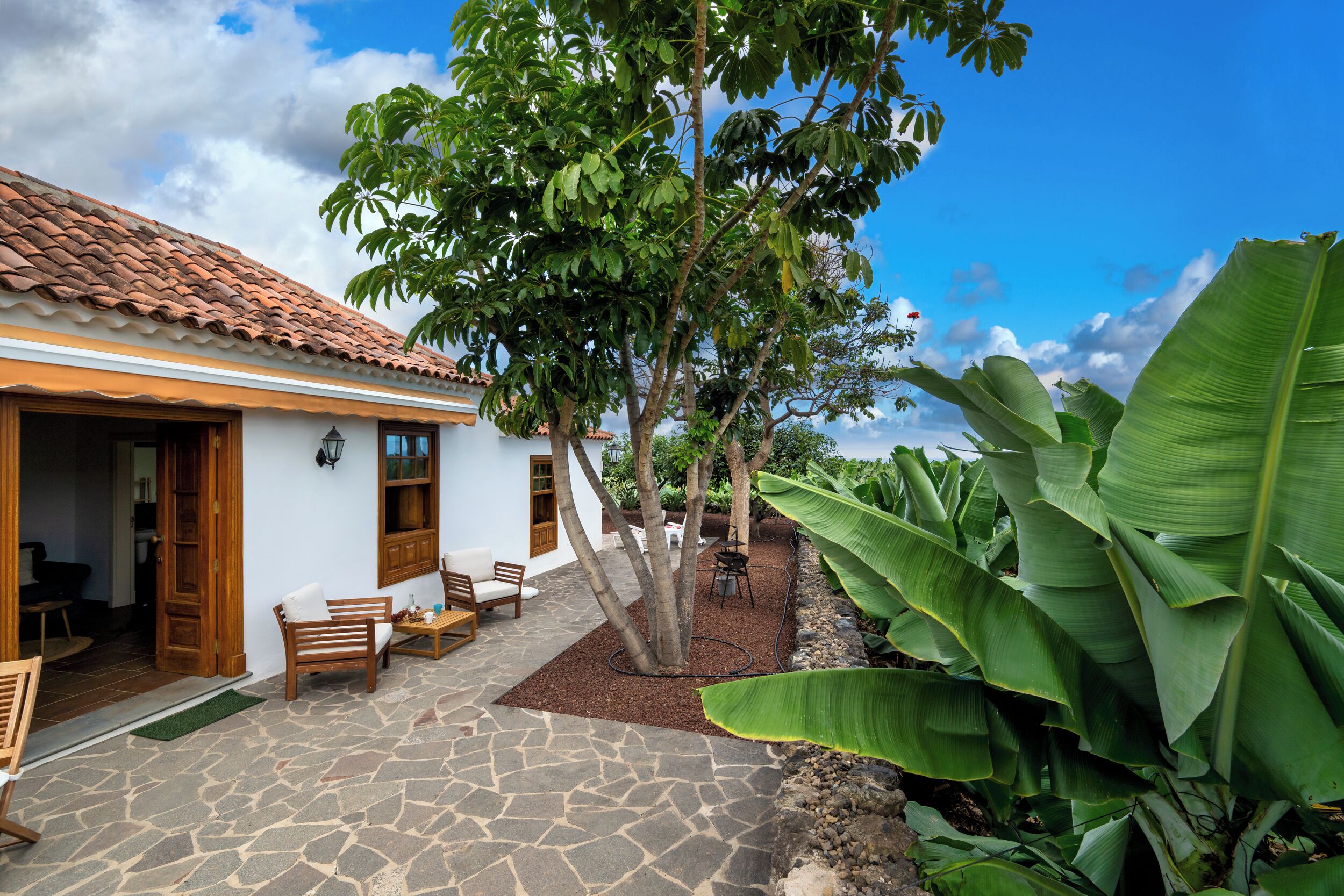 Un patio con techo de tejas, suelo de piedra, muebles de madera y plantas verdes grandes.