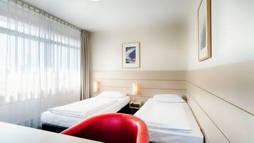A hotel room with two single beds, a red chair, a desk, and a large window with sheer curtains.