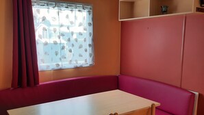A room with a wooden table and chairs, a red cushioned bench, a window with a leaf-patterned curtain, and a shelf with a bird figure.