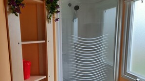 A shower with a glass door, a red bucket on a shelf, and a window with blinds.