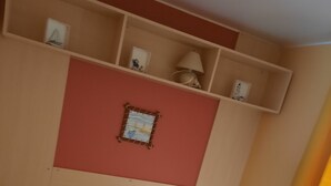 A hotel room with two single beds, a small table, and a shelf with personal items.