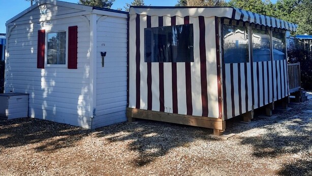 Two small, single-story cabins with striped awnings, one with a red door and the other with a black door, set on a gravel ground with trees in the background.
