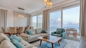 A spacious living room with a sofa, armchairs, a coffee table, and a dining area with chairs and a table. Large windows offer a view of the sea.