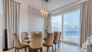 A dining area with a round table and six chairs, a sofa, and a large window with a view of the sea.