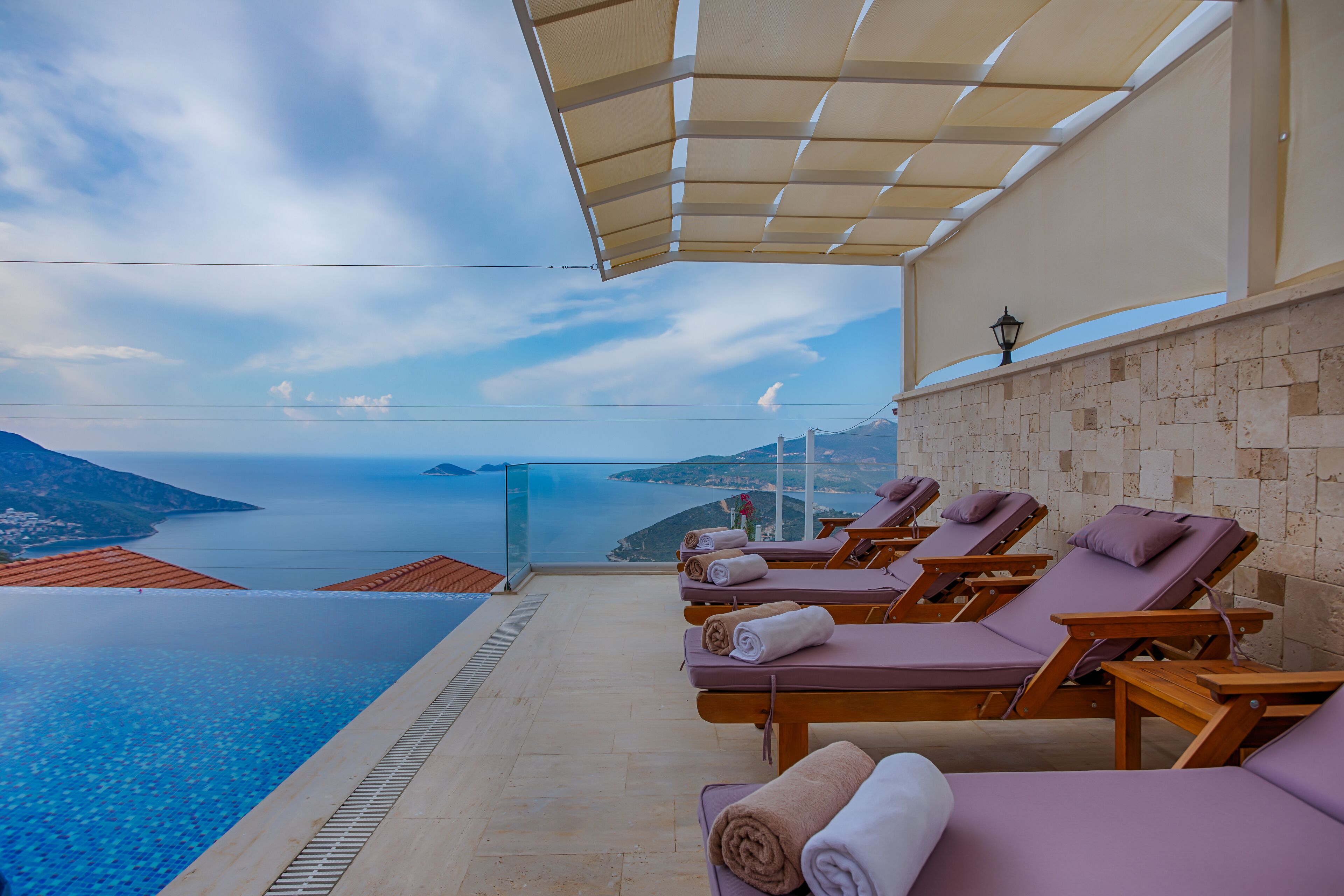 An infinity pool with a view of the sea, sun loungers with towels, and a stone wall.