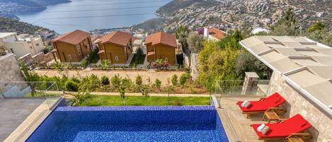 A swimming pool with blue mosaic tiles, red lounge chairs, and a view of a coastal town with houses and hills.
