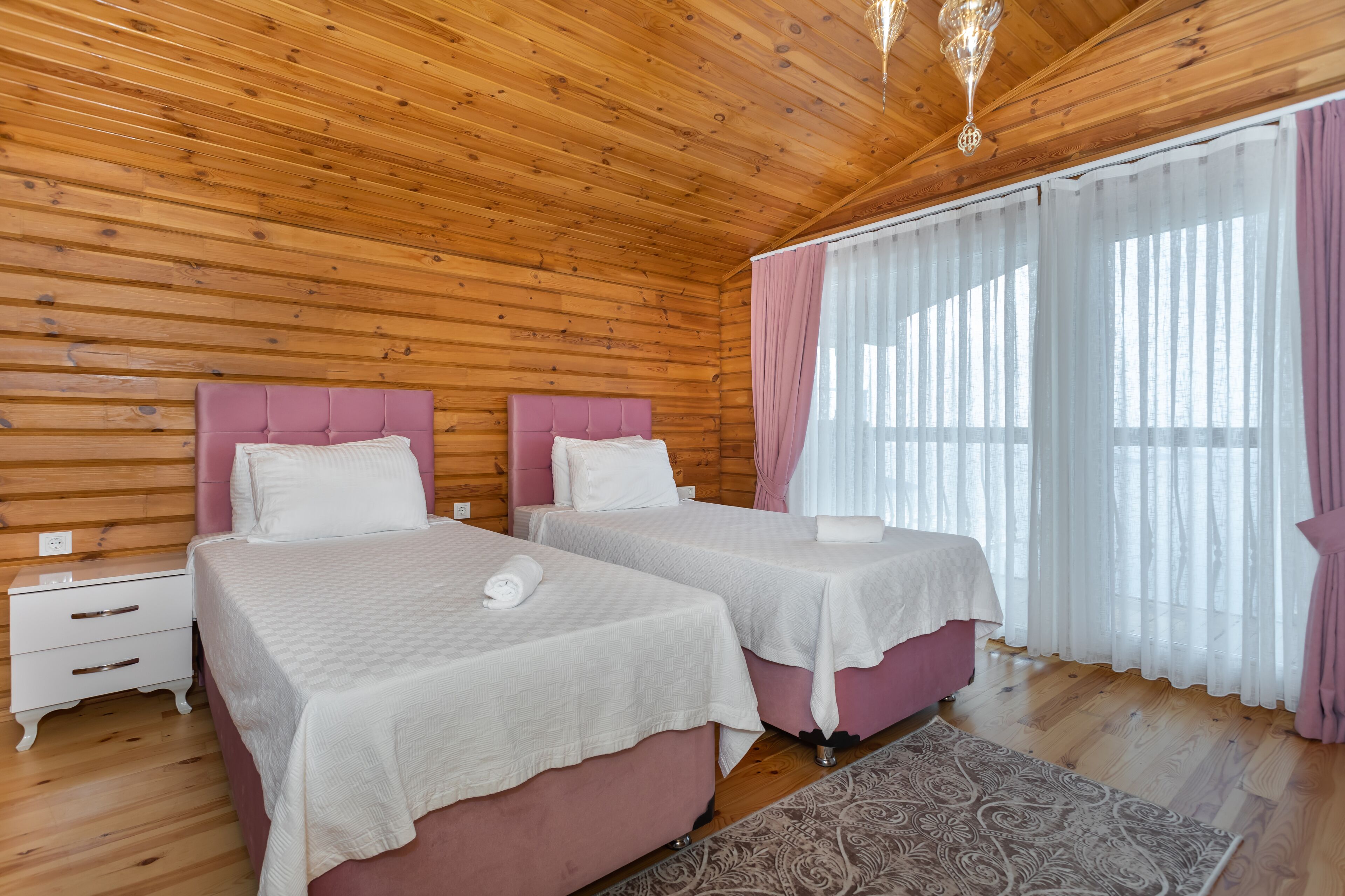 A wooden-paneled room with two beds, a nightstand, and a window with sheer curtains.