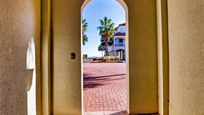 An arched doorway leading to a paved area with palm trees and a building in the background. - Southern Star - Across Street From Beach, Bay Views. Community Pool! (St George Island)
