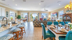 A spacious kitchen with a large island, a dining area with a wooden table set for six, and a living area with a patterned sofa and teal chairs. - Southern Star - Across Street From Beach, Bay Views. Community Pool! (St George Island)