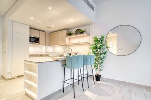 A modern kitchen with white cabinetry, a central island with bar stools, a potted plant, and a round mirror on the wall. - Opulent 1BR at Belgravia Tower JVC by Deluxe Holiday Homes (Dubai)