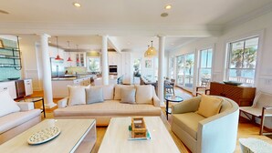 Amplia sala de estar con un sofá beige grande, un sillón más pequeño y una mesa de centro. Se observa una cocina al fondo con armarios blancos y lámparas colgantes rojas.