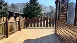 A wooden deck with a wooden fence, a building with large windows, and trees in the background. - Cabin 38 -  Retreat Group (Aviemore)