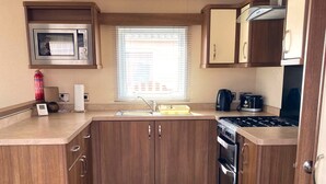 A compact kitchen with wooden cabinets, a microwave, a stove, and a window with blinds. - Cabin 38 -  Retreat Group (Aviemore)