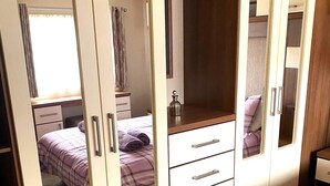 A bedroom with a large wardrobe, a bed with a purple checkered bedspread, and a small bedside table with a lamp. - Cabin 38 -  Retreat Group (Aviemore)