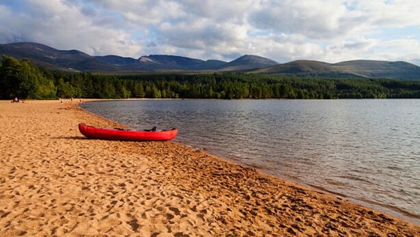A sandy beach with a red kayak and a calm lake, surrounded by mountains and dense forest. - Cabin 38 -  Retreat Group (Aviemore)