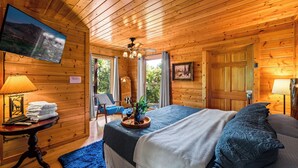 A wooden cabin room with a bed, a TV mounted on the wall, a table with towels and a bottle, and a chair by the window. - Mountain View! Cabin, 6BR, Yard, Firepit, GameRoom (Sevierville)