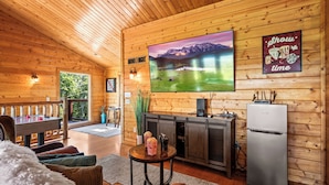 A cozy cabin interior with a wooden ceiling, a large flat-screen TV, a dining table, and a refrigerator. - Mountain View! Cabin, 6BR, Yard, Firepit, GameRoom (Sevierville)