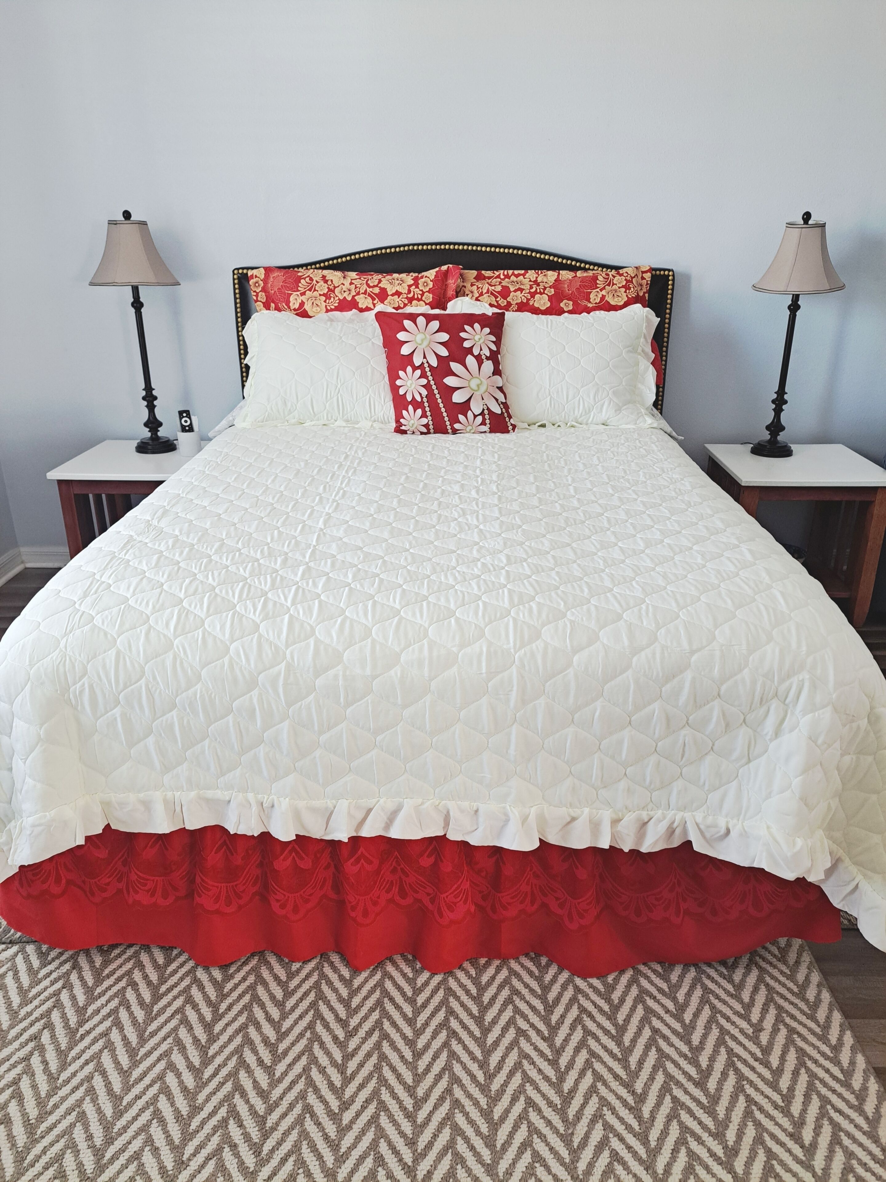 A bed with a white quilt and red floral pillow, flanked by two bedside tables with lamps, against a light blue wall.