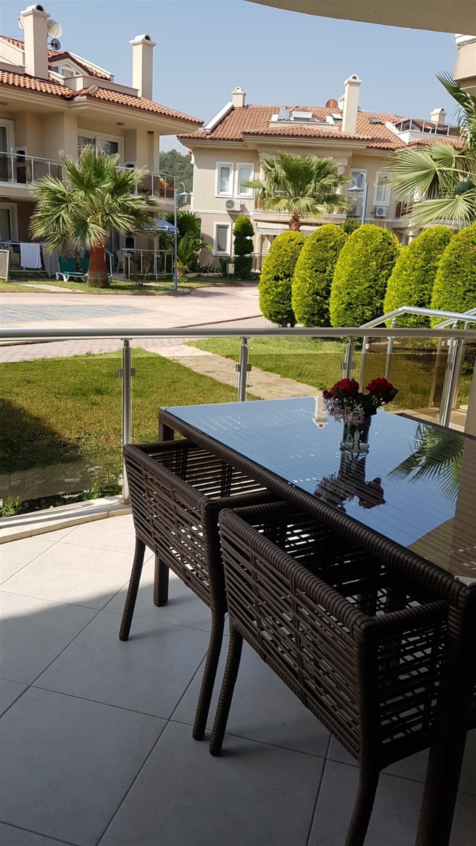 Ein Tisch mit Glasplatte und Korbstühle auf einem Balkon mit Blick auf einen gepflegten Garten und angrenzende Häuser.