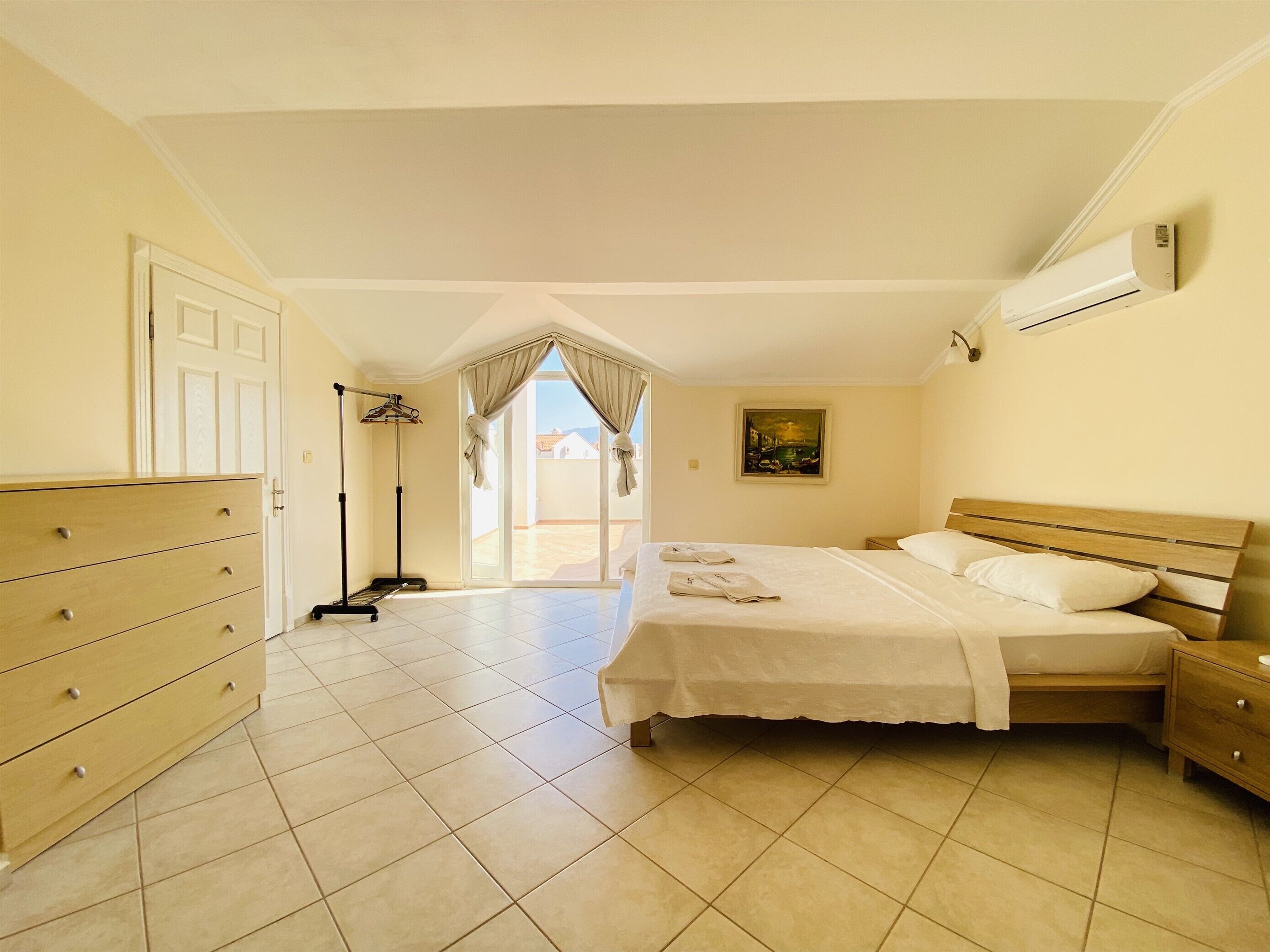 A hotel room with a bed, a dresser, a painting, and an air conditioning unit.