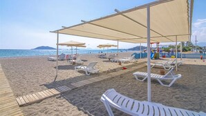 En strand med solstolar och parasoller under ett tak, en träpromenad och en lekplats i bakgrunden.
