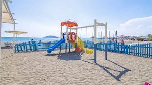 En lekplats vid stranden med en rutschkana och gungor, omgiven av ett blått staket och parasoller.