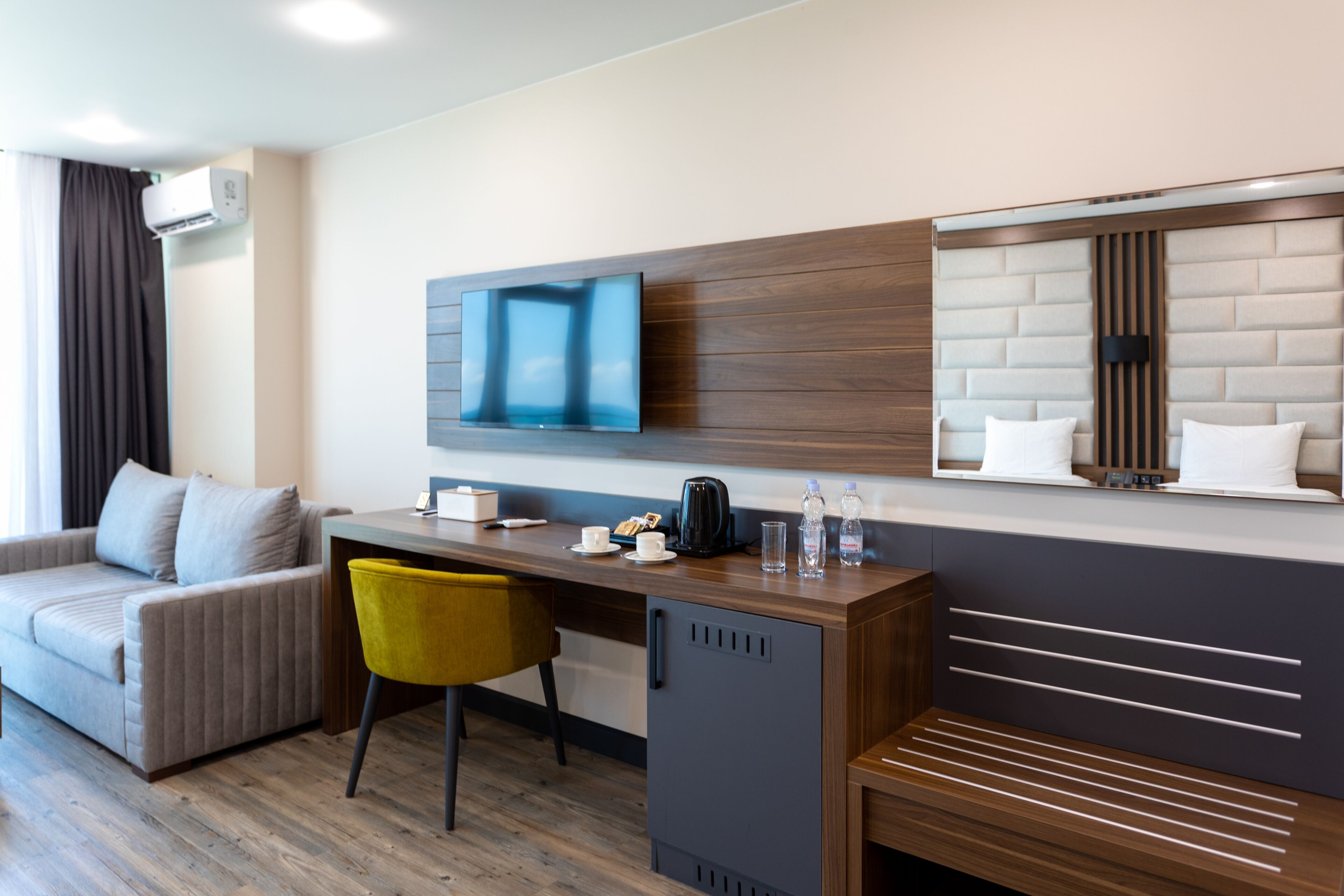 Una habitación de hotel moderna con sofá, escritorio, silla, televisor, mini refrigerador y estante empotrado con espejo.