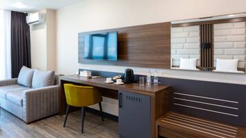 Una habitación de hotel moderna con sofá, escritorio, silla, televisor, mini refrigerador y estante empotrado con espejo.