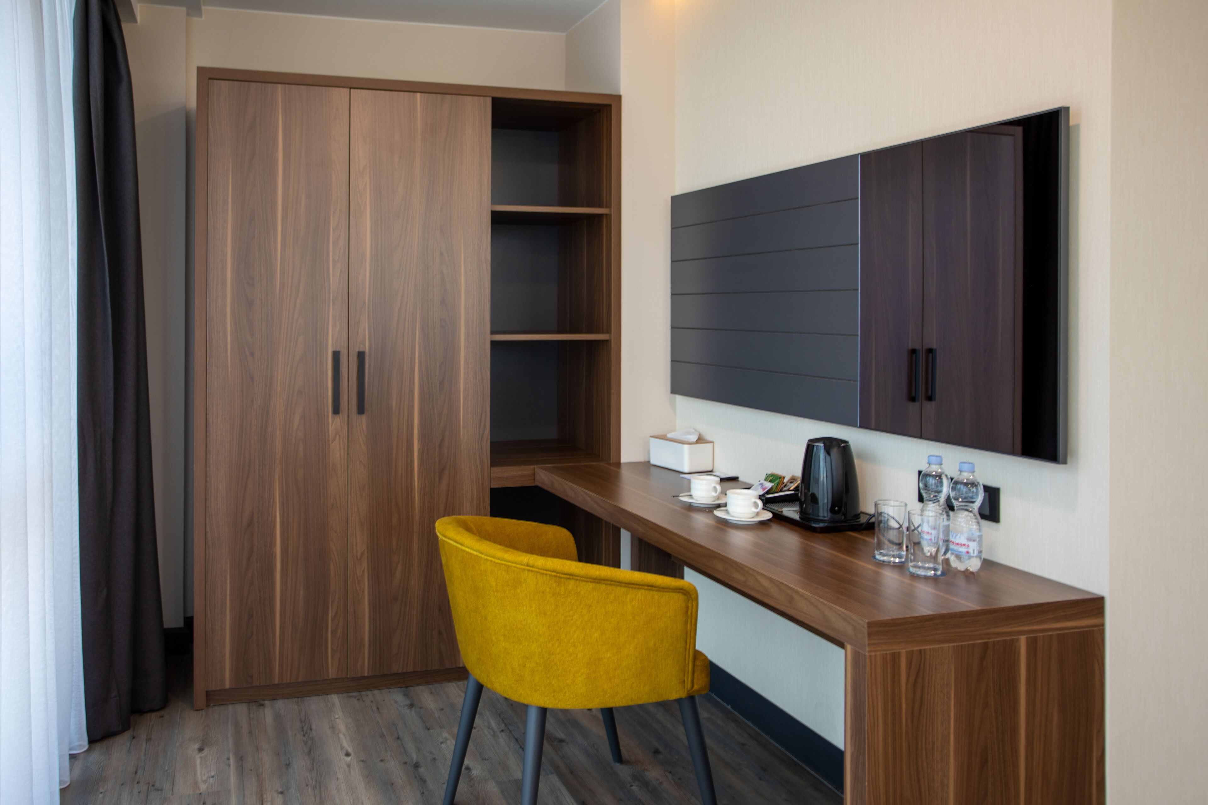A compact room with a wooden wardrobe, a yellow chair, a small table with a kettle, cups, and bottles.