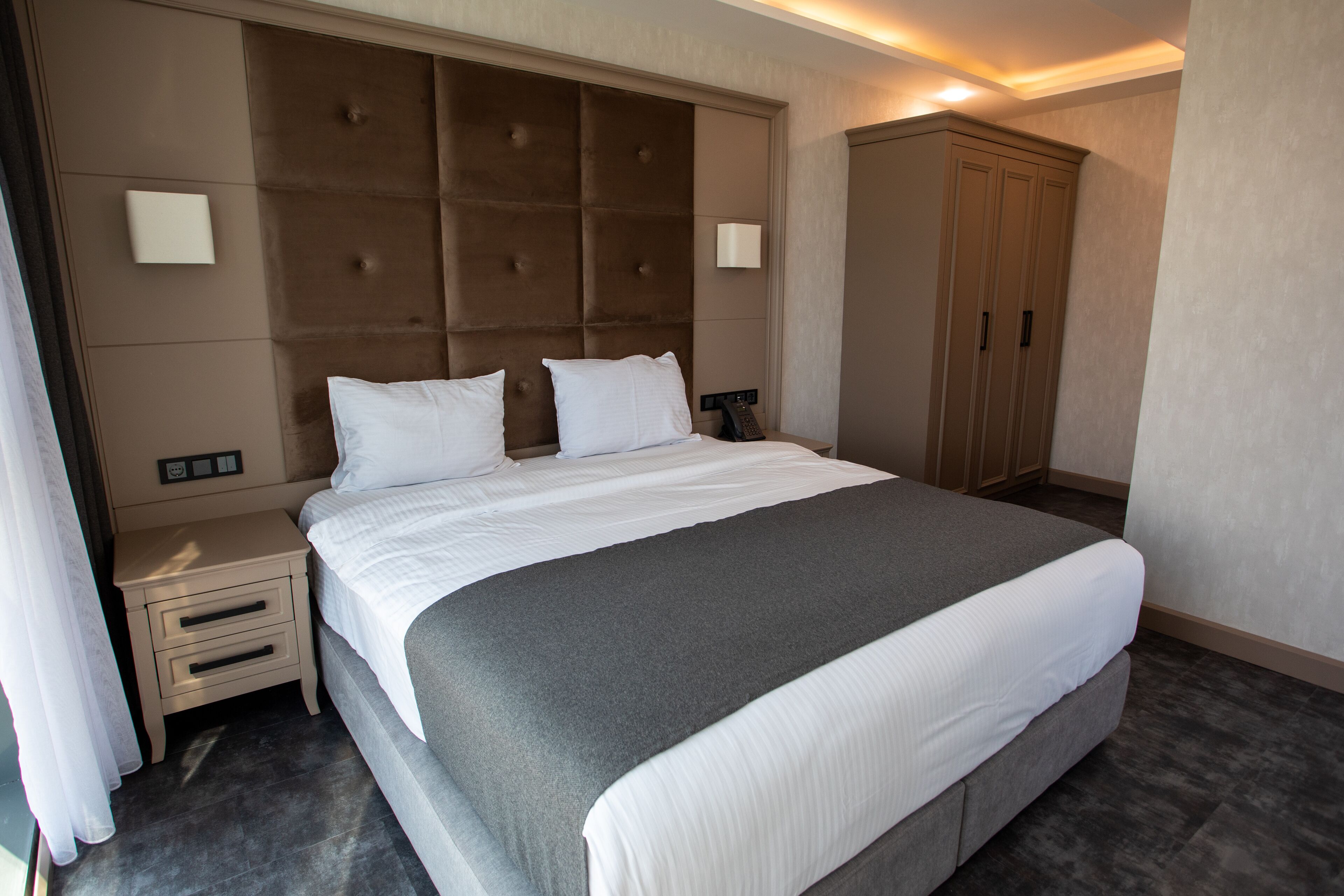 A modern hotel room with a large bed, a nightstand, a wardrobe, and a built-in headboard.