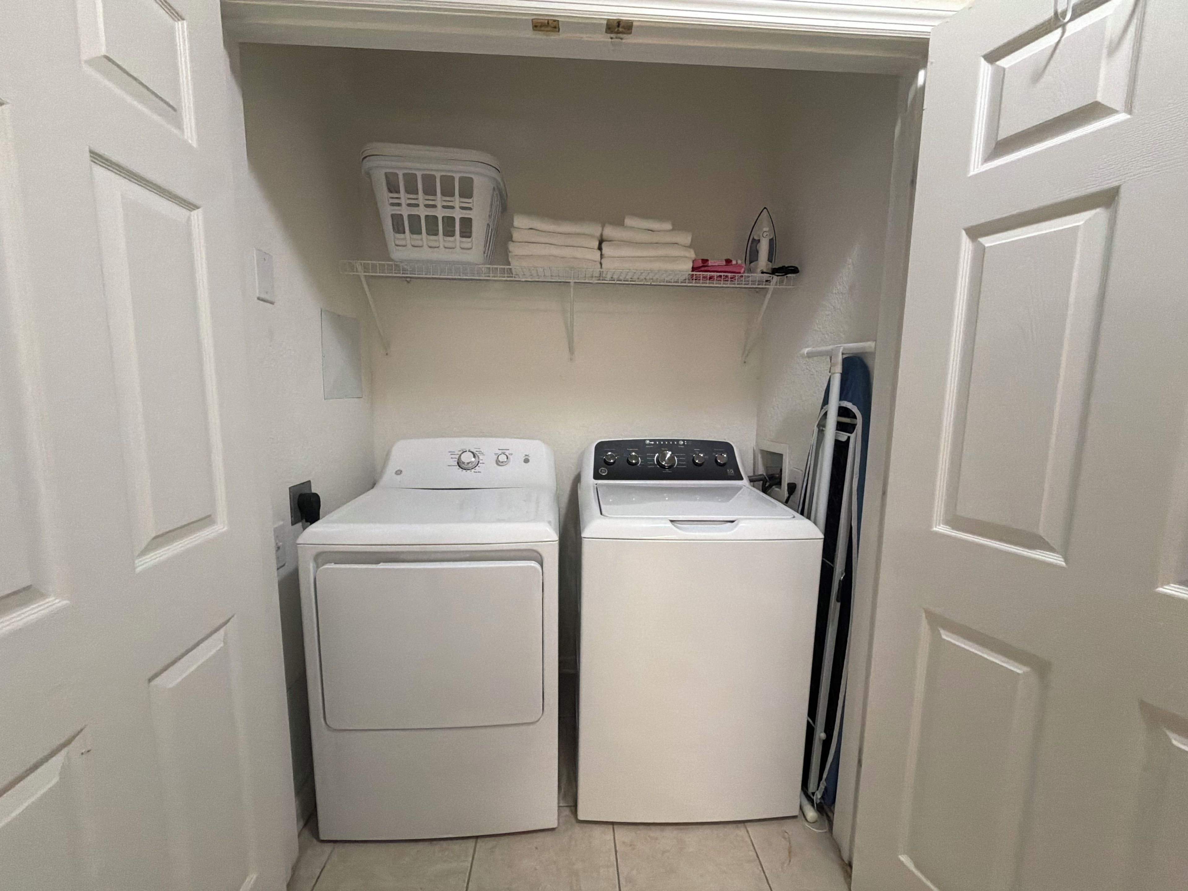 A laundry room with two white front-loading washing machines and a shelf above containing folded towels and a laundry basket.
