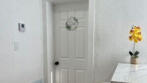 Una puerta blanca con decoración geométrica, una pequeña planta en maceta y una mesa blanca con una silla en una habitación con paredes blancas y suelo de madera.
