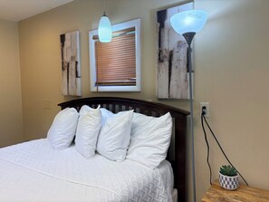 A bedroom with a bed, white pillows, a nightstand with a plant, and wall art.
