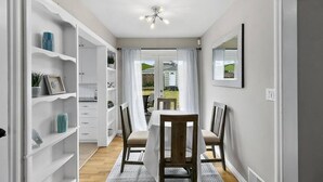 Een eethoek met een tafel en stoelen, een witte wandplank met decoratieve objecten, en een uitzicht door een glazen deur naar buiten.