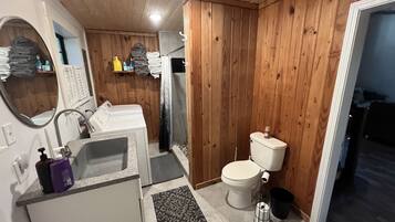 Et kompakt badeværelse med trævægge, et hvidt toilet, en vask med spejl og et lille bruseområde.