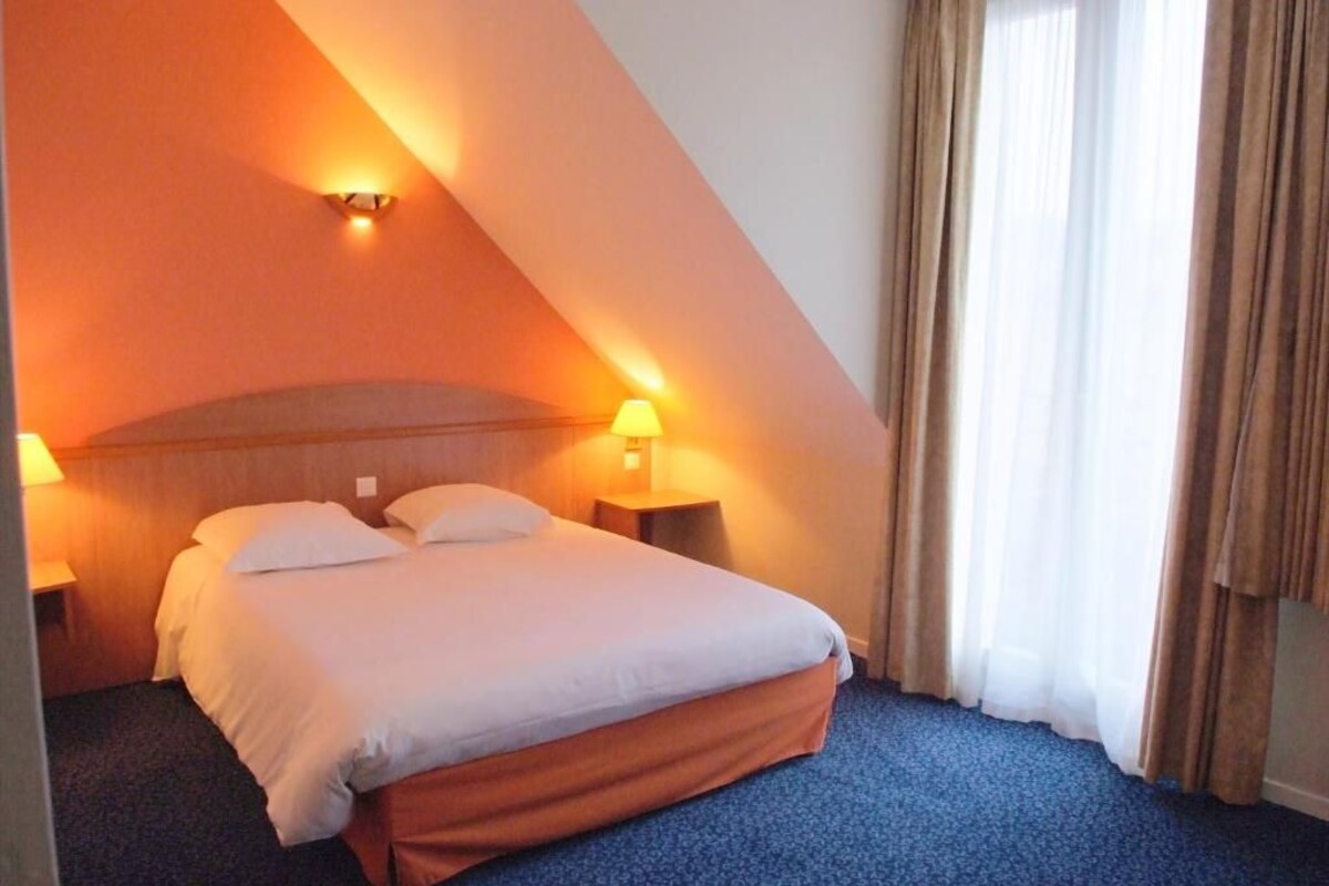 Une chambre d’hôtel avec un lit, deux lampes de chevet, une tête de lit en bois et une fenêtre avec des rideaux.