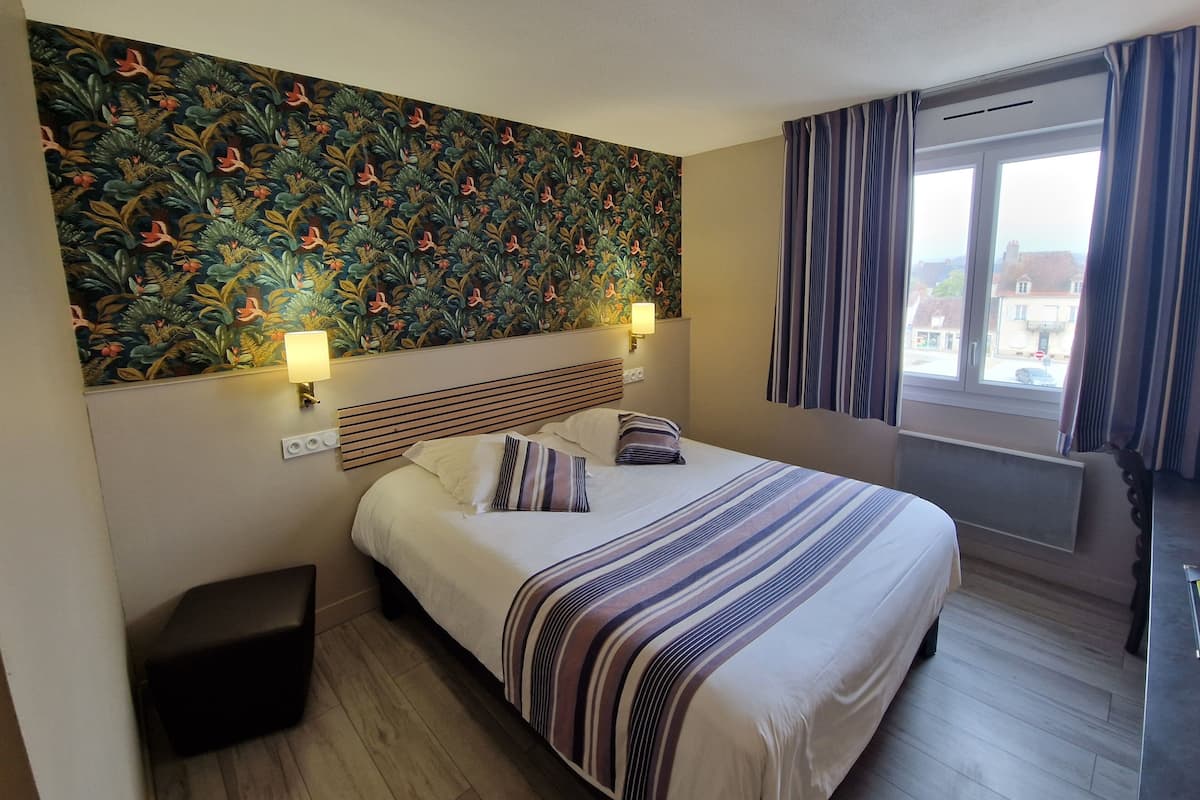 Hotelkamer met groot bed, hoofdbord met horizontale latten, wand met bloemenpatroon, raam met gordijnen, en klein nachtkastje met lamp.