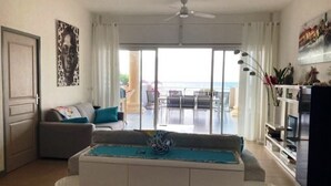 Een woonkamer met een bank, een salontafel, een tv-meubel en uitzicht op de oceaan door grote ramen.