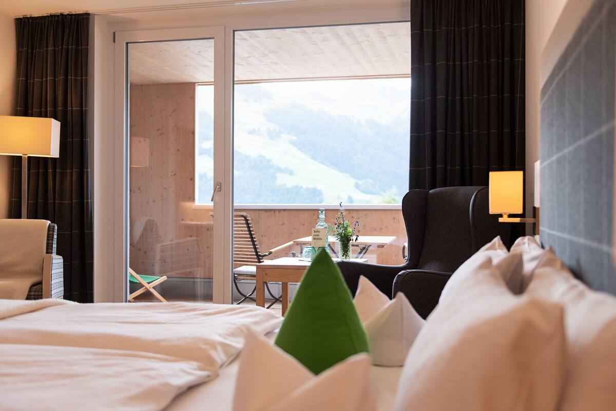 Quarto de hotel com cama, uma cadeira, uma mesa e vista para montanhas através de uma grande janela.