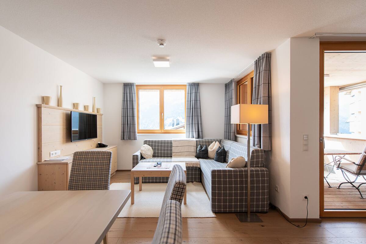 Uma sala de estar moderna com sofá, televisão na parede, mesa de jantar e vista para o exterior através de grandes janelas.