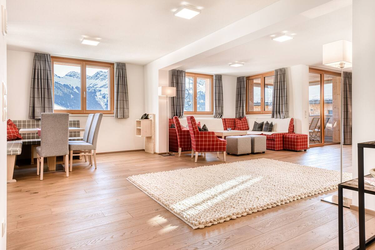 Uma sala de estar ampla com piso de madeira, um tapete grande e um espaço de estar com cadeiras vermelhas e cinzas.