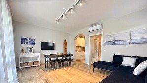 Een moderne woonkamer met een donkere bank, een eethoek met een houten tafel en stoelen, en een keuken op de achtergrond.